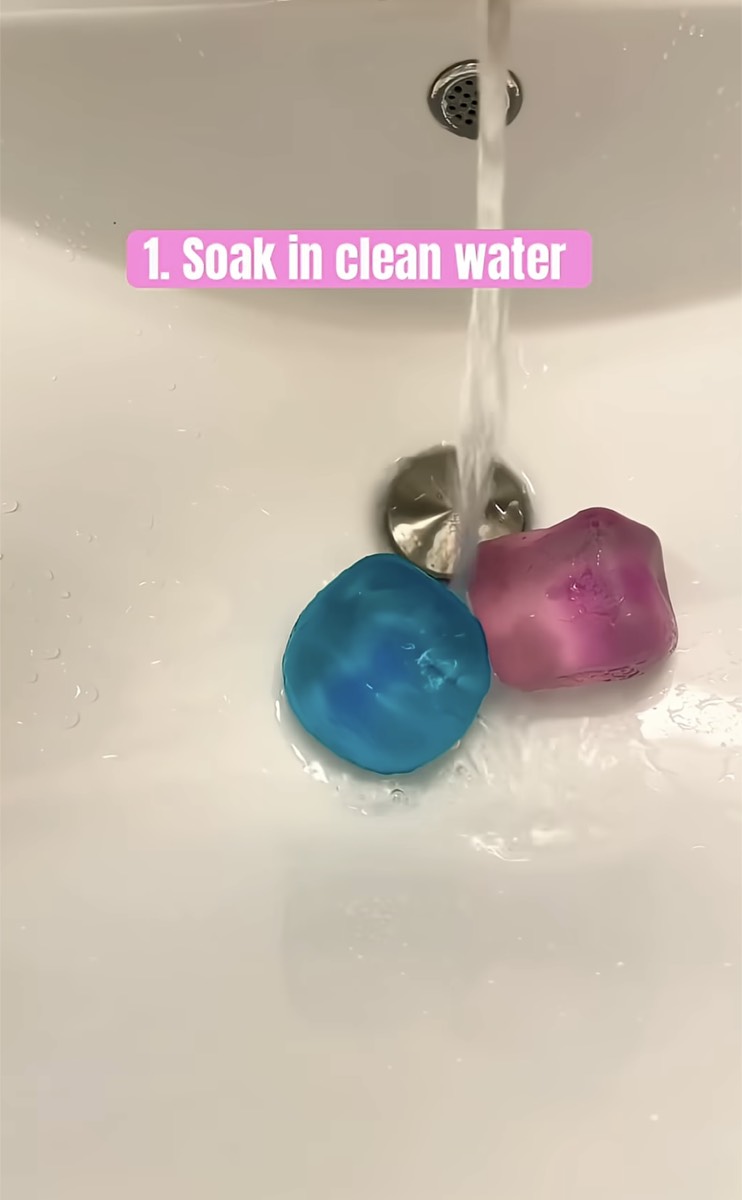 Two clear NeeDoh cubes under running water in a white sink