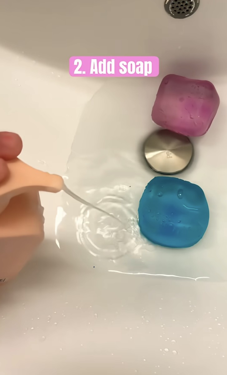 A finger adding a drop of soap to the water beside two NeeDoh cubes
