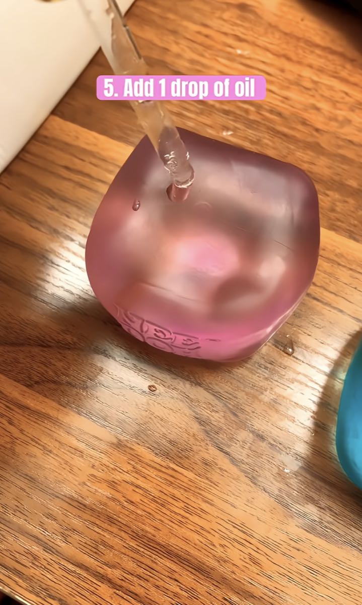 A dropper putting one drop of oil onto a clear purple NeeDoh cube on a wooden table