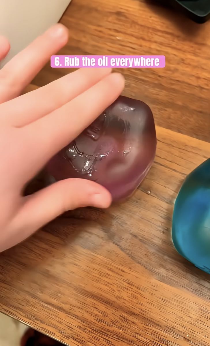 Fingers spreading a thin film of oil across a purple NeeDoh cube